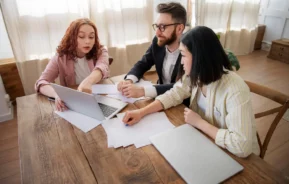 Imagem de duas mulheres e um homem sentados em cadeiras ao redor de uma mesa olhando informações na tela de um computador para ilustrar matéria sobre partilha de imóvel financiado