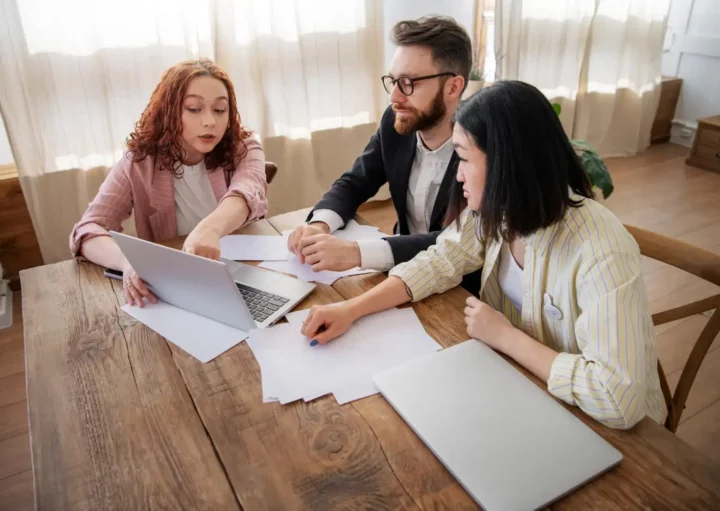 Imagem de duas mulheres e um homem sentados em cadeiras ao redor de uma mesa olhando informações na tela de um computador para ilustrar matéria sobre partilha de imóvel financiado