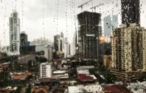 Imagem de janela embaçada e com gotículas de chuva em primeiro plano e de prédios e casas residenciais em segundo plano para ilustrar matéria sobre qual a cidade que mais chove em Santa Catarina