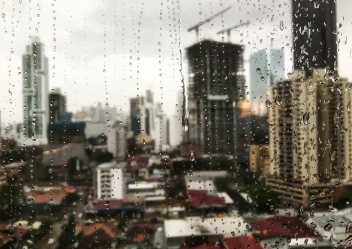 Imagem de janela embaçada e com gotículas de chuva em primeiro plano e de prédios e casas residenciais em segundo plano para ilustrar matéria sobre qual a cidade que mais chove em Santa Catarina