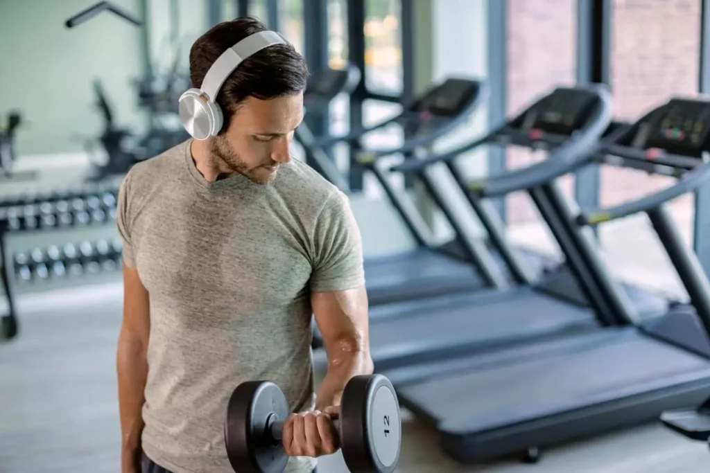 Imagem de um homem em uma academia com fone de ouvido e camisa suada enquanto levanta um peso de mão na frente de algumas esteiras para ilustrar matéria sobre academia de condomínio