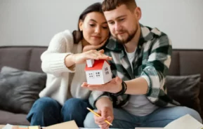 Imagem de um casal composto por um homem e uma mulher sentados no sofá da sala olhando para uma casa em tamanho miniatura que está na mão do homem enquanto anotam e planejam em cadernos que estão em uma mesinha de centro para ilustrar matéria sobre o cheque moradia