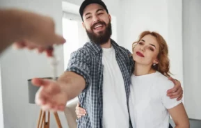 Imagem de um casal formado por um homem e uma mulher se abraçando enquanto recebem a chave de um imóvel de uma terceira pessoa para ilustrar matéria sobre como comprar um apartamento ganhando um salário mínimo