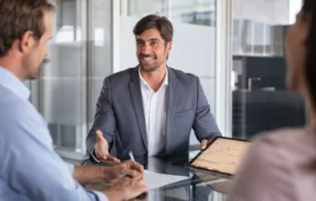 Imagem que ilustra componente de Imovelweb mostra um corretor sentado em frente à dois clientes conversando