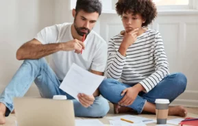 Imagem de um casal composto por um homem e uma mulher sentados no chão de uma casa analisando papéis que estão no chão ao lado de um notebook para ilustrar matéria sobre desistência de compra de imóvel entre particulares