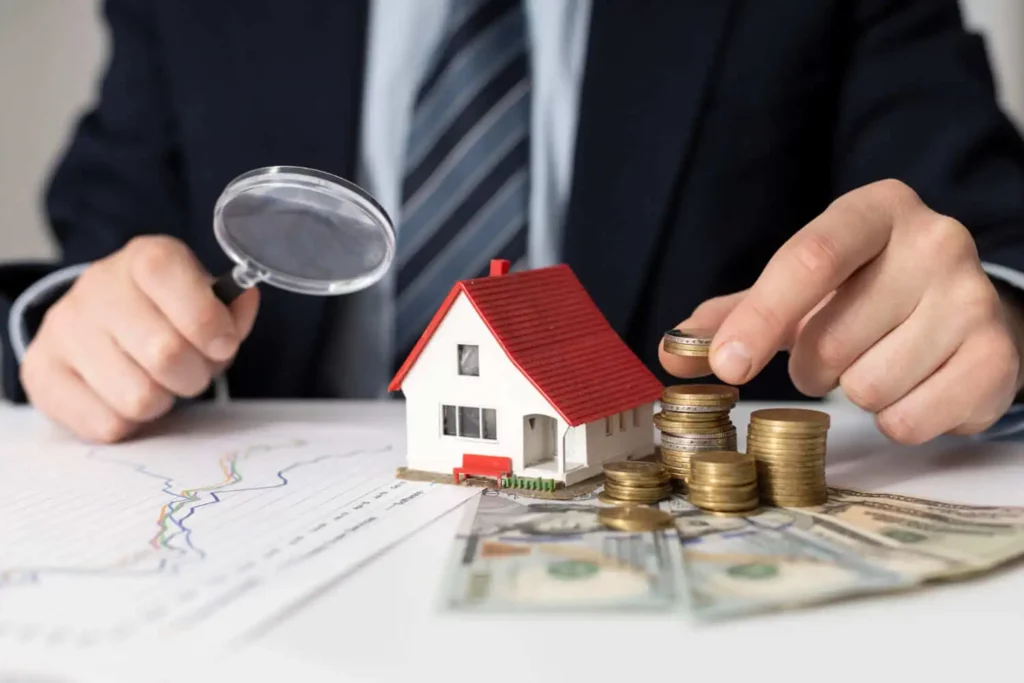 Imagem de um homem segurando uma lupa enquanto empilha algumas moedas que estão em cima de uma mesa ao lado de uma casa em tamanho miniatura para ilustrar matéria sobre financiamento imobiliário, como calcular?