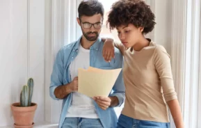 Imagem de um homem e uma mulher um do lado do outro encostados na parede de uma casa olhando para um papel que está na mão do homem para ilustrar matéria que responde a pergunta: imóvel de herança, o cônjuge tem direito?