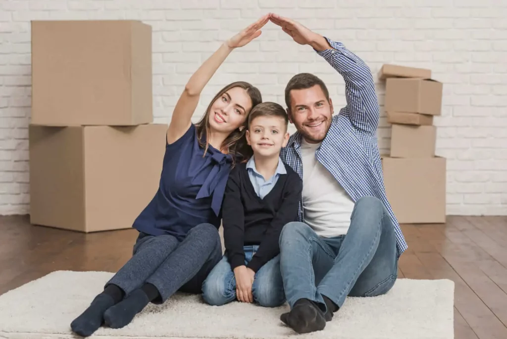 Imagem de uma família composta por um homem, mulher e criança sentados no chão formando uma casa com as mãos em um cômodo com caixas de mudança para ilustrar matéria sobre o que é seguro FGHab