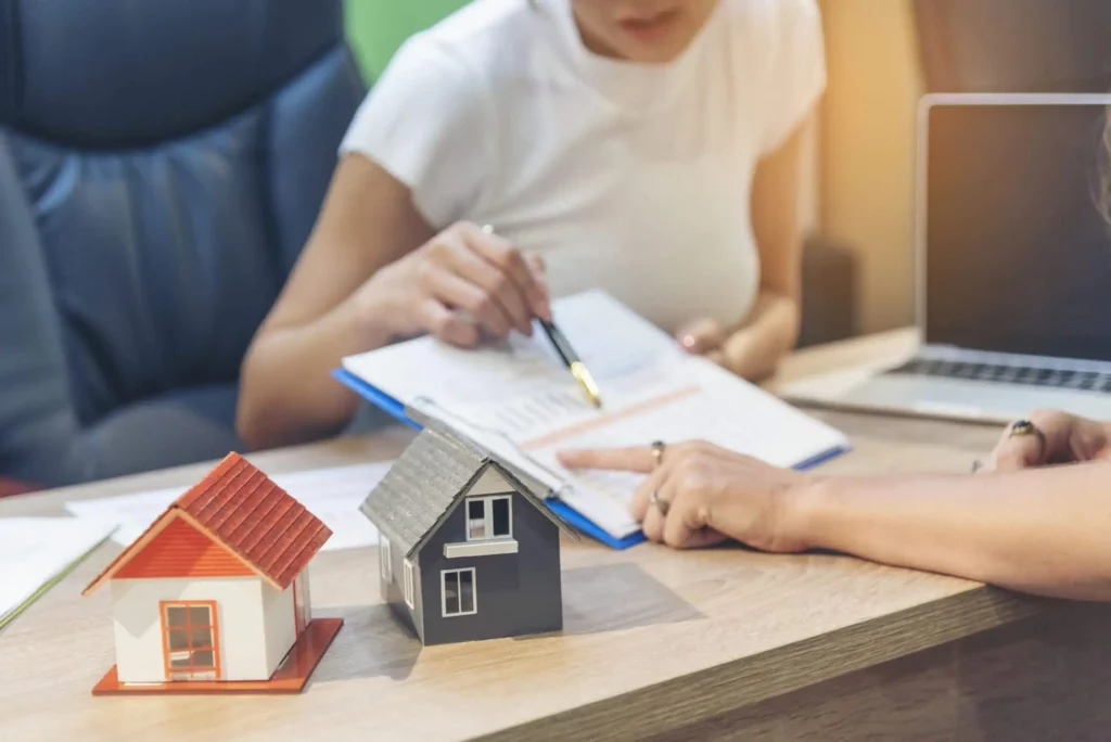 Imagem de uma pessoa analisando documentos com outra pessoa em uma mesa, com duas pequenas casas modelo e um laptop por perto