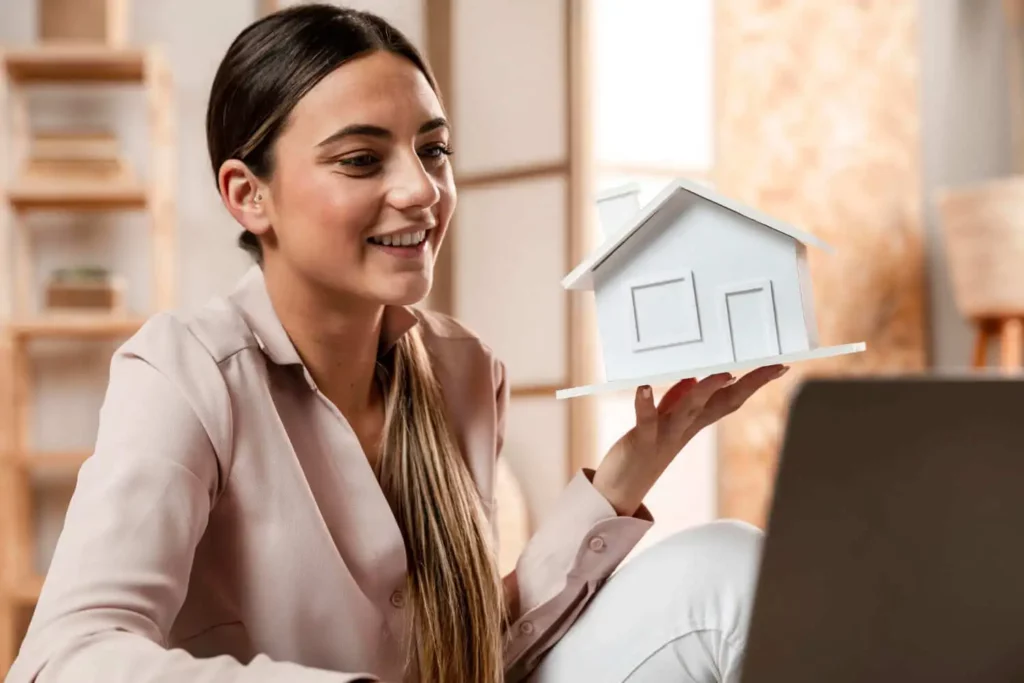 Imagem de uma mulher segurando uma casa de madeira em tamanho miniatura na cor branca enquanto olha para a tela de um notebook com um sorriso no rosto para ilustrar matéria sobre como calcular a liquidez de um imóvel