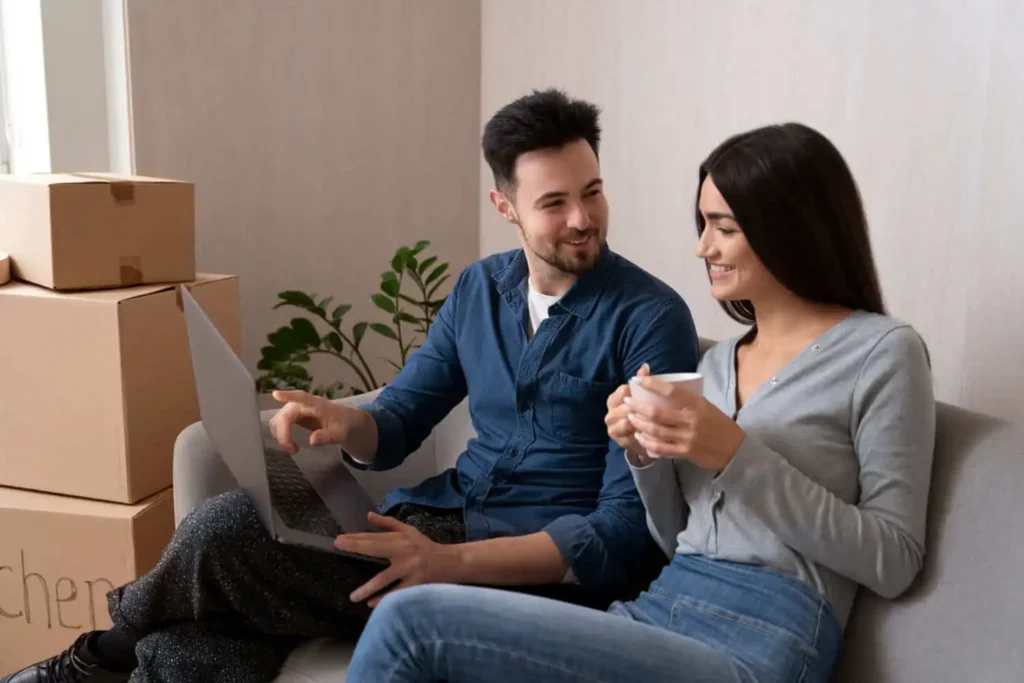 Imagem de um casal formado por um homem e uma mulher sentados em um sofá ao lado de caixas de papelão olhando para a tela de um notebook para ilustrar matéria sobre como ganhar dinheiro com imóvel