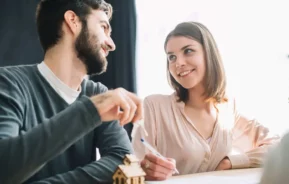 Imagem de um casal formado por um homem e uma mulher se olhando enquanto assinam um documento que está em cima de uma mesa ao lado de uma casa de maneira em tamanho miniatura para ilustrar matéria sobre como investir em imóveis com pouco dinheiro