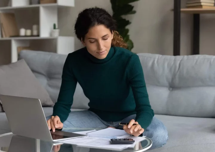 Imagem de uma mulher sentada em um sofá, trabalhando em uma mesa baixa com um laptop, papéis, uma caneta e uma calculadora. Ela parece concentrada, analisando documentos e fazendo anotações sobre o custo efetivo total em um ambiente aconchegante de sala de estar