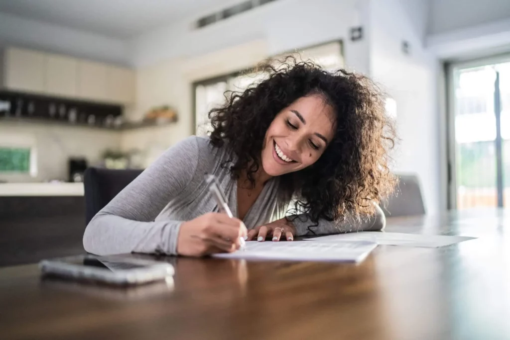 Imagem de uma mulher de cabelos cacheados sorrindo enquanto escreve em uma mesa de madeira em uma casa moderna e iluminada. Um smartphone e documentos estão sobre a mesa, e a cozinha pode ser vista ao fundo