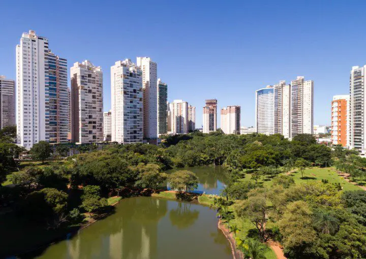 Imagem do Parque Flamboyant, no bairro Jardim Goiás, mostra uma paisagem urbana com arranha-céus altos e modernos atrás de um parque verde exuberante com árvores e um lago sob um céu azul claro para ilustrar matéria sobre os bairros nobres de Goiânia.