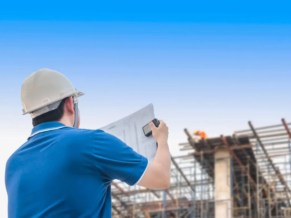 Imagem de um homem vestido com uma camisa azul e chapéu de proteção para construção analisando a planta de um prédio em construção em frente à obra para ilustrar matéria sobre preço para construir por metro quadrado