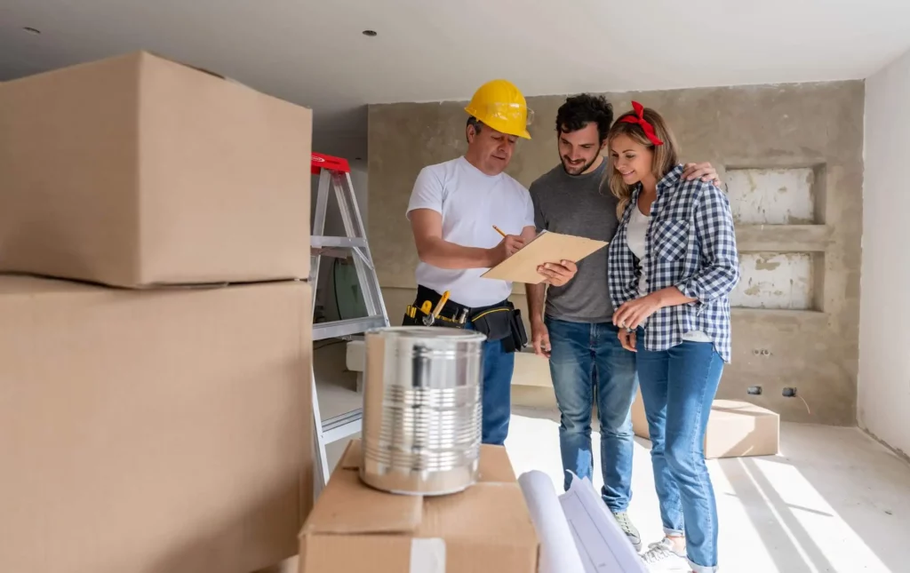 Imagem de um trabalhador da construção civil com um capacete amarelo mostra uma prancheta a um casal sorridente dentro de um quarto parcialmente reformado, cercado por caixas de mudança e plantas enroladas