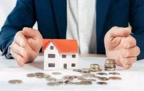 Imagem da silhueta de um homem sentado em uma mesa com as mãos entre uma casa em tamanho miniatura e algumas moedas que estão em cima de uma mesa para ilustrar matéria sobre valor de liquidez de imóvel