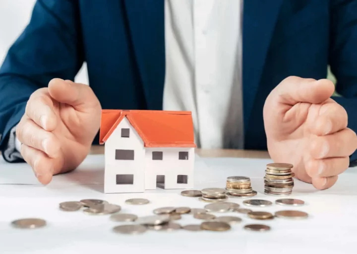 Imagem da silhueta de um homem sentado em uma mesa com as mãos entre uma casa em tamanho miniatura e algumas moedas que estão em cima de uma mesa para ilustrar matéria sobre valor de liquidez de imóvel