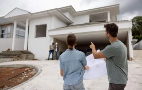 Imagem de um homem e uma mulher do lado de fora de uma casa branca moderna em construção, segurando plantas e discutindo o valor do metrô quadrado construído, enquanto dois trabalhadores conversam perto da garagem