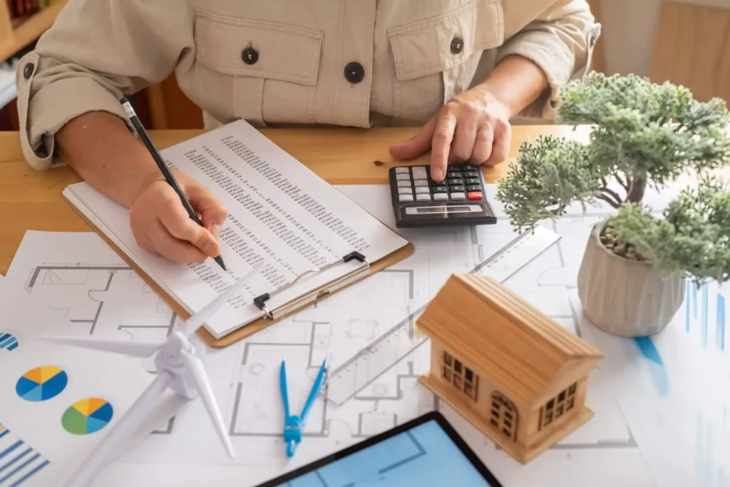 Imagem de uma pessoa escrevendo em uma prancheta com dados financeiros, cercada por plantas de casas, gráficos, uma casa modelo, um modelo de turbina eólica e um vaso de planta sobre a mesa