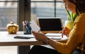 Imagem que ilustra matéria sobre como atualizar o valor do imóvel no imposto de renda mostra uma moça vendo um papel e anotando sentada em uma mesa em frente à um computador