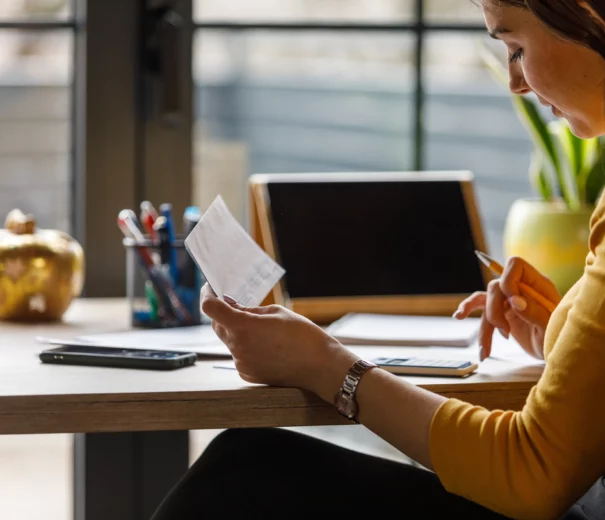 Imagem que ilustra matéria sobre como atualizar o valor do imóvel no imposto de renda mostra uma moça vendo um papel e anotando sentada em uma mesa em frente à um computador