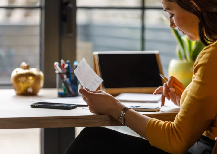 Imagem que ilustra matéria sobre como atualizar o valor do imóvel no imposto de renda mostra uma moça vendo um papel e anotando sentada em uma mesa em frente à um computador