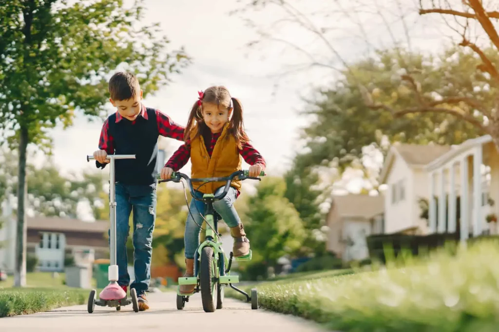 Imagem de duas crianças brincando na rua para ilustrar matéria sobre um bairro em Nilópolis. Enquanto um menino está de patinete, uma menina ao lado está pedalando uma bicicleta em um bairro residencial