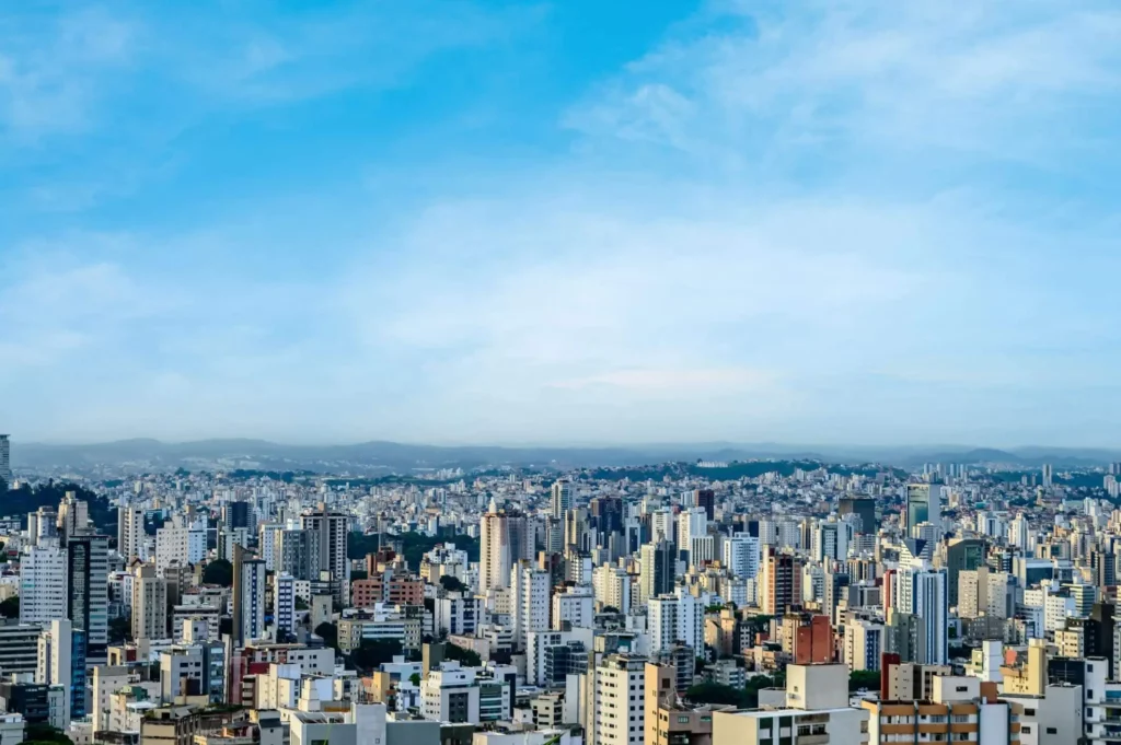 Imagem da vista panorâmica de Belo Horizonte mostra prédios altos sob um céu azul brilhante e nuvens dispersas. A paisagem urbana se estende até as colinas distantes no horizonte