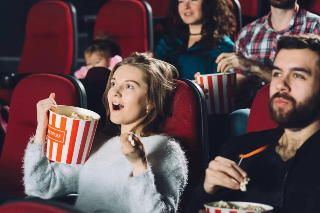 Imagem de uma mulher com expressão de surpresa sentada em uma poltrona de uma sala de cinema ao lado de um homem segurando um balde de pipoca para ilustrar matéria sobre cinema em Uberlândia