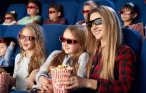 Imagem de uma mulher e várias crianças sentadas nas poltronas de uma sala de cinema assistindo a um filme com óculos 3D no rosto e balde de pipoca nas mãos para ilustrar matéria sobre os cinemas em Uberlândia