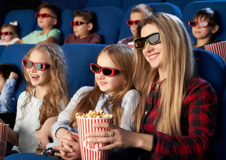 Imagem de uma mulher e várias crianças sentadas nas poltronas de uma sala de cinema assistindo a um filme com óculos 3D no rosto e balde de pipoca nas mãos para ilustrar matéria sobre os cinemas em Uberlândia