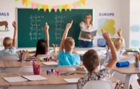 Imagem de uma sala de aula cheia de crianças com braço levantado e uma professora com um livro na mão em frente a uma lousa branca para ilustrar matéria sobre escola em Nova Iguaçu