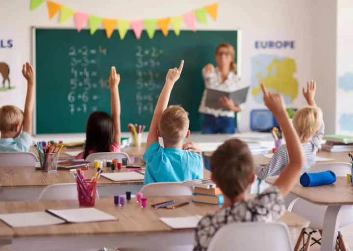 Imagem de uma sala de aula cheia de crianças com braço levantado e uma professora com um livro na mão em frente a uma lousa branca para ilustrar matéria sobre escola em Nova Iguaçu