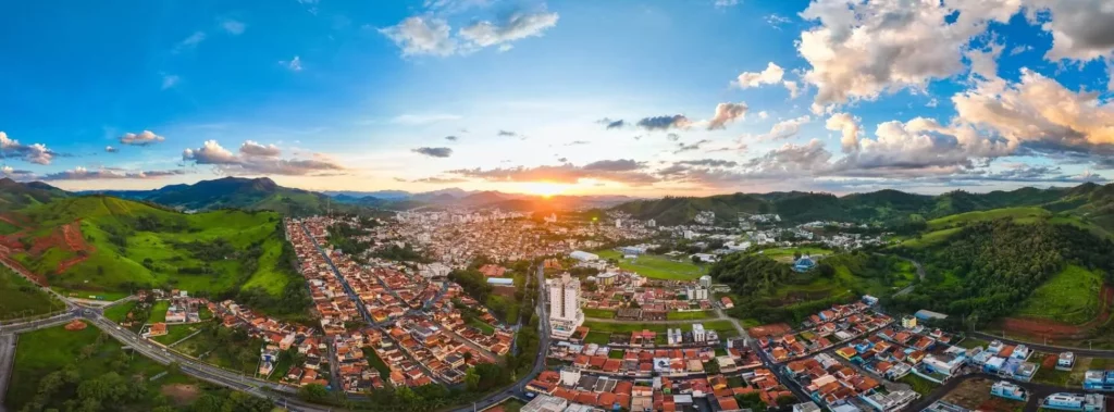 Imagem da vista aérea da cidade de Itajubá, em Minas Gerais, mostra telhado de casas, avenidas e vegetação em um dia de céu azul com nuvens claras