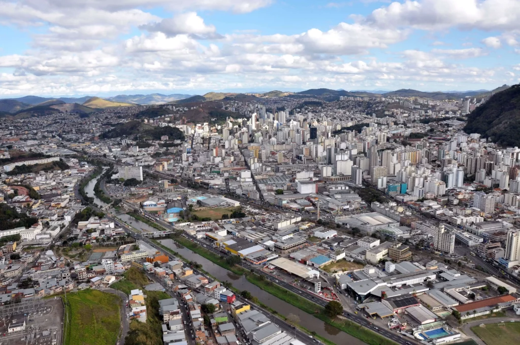 Imagem da vista aérea de Juiz de Fora, em Minas Gerais, mostra prédios altos, áreas industriais e um rio correndo pelo centro, cercada por colinas e montanhas verdes sob um céu parcialmente nublado