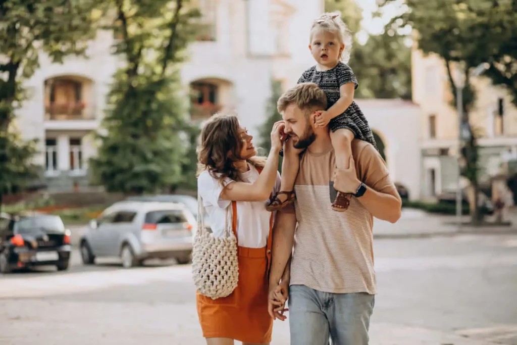 Imagem de uma família formada por um homem, uma mulher e uma criança que está em cima dos ombros do homem passeando pelas ruas de uma cidade para ilustrar matéria sobre os melhores bairros em Vinhedo para morar