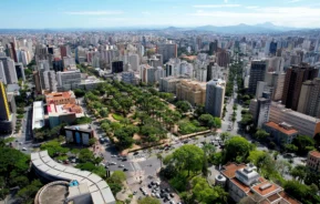 Imagem da vista aérea de Belo Horizonte mostra prédios altos ao redor de um parque central repleto de árvores e pistas de caminhada. Ruas movimentadas, carros e vegetação são visíveis sob um céu azul claro