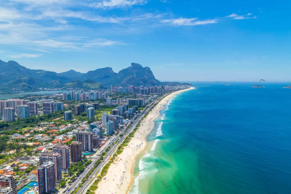 Imagem aérea da praia da Barra mostra mar, faixa de areia, prédios e vegetação da área para ilustrar matéria sobre as melhores praias da Barra da Tijuca