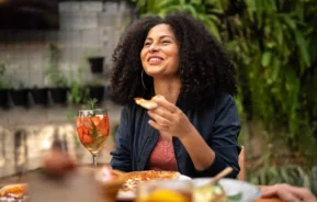 Imagem de uma mulher adulta sentada em uma mesa com algumas comidas em cima sorrindo, conversando com alguns amigos em um restaurante e comendo para ilustrar matéria sobre os melhores restaurantes em São Paulo