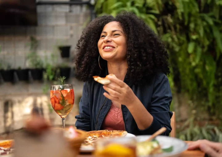 Imagem de uma mulher adulta sentada em uma mesa com algumas comidas em cima sorrindo, conversando com alguns amigos em um restaurante e comendo para ilustrar matéria sobre os melhores restaurantes em São Paulo