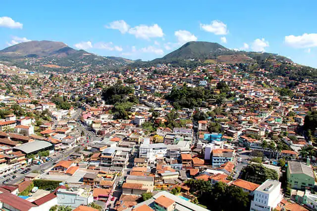 Imagem da vista aérea de Nova Lima, em Minas Gerais, mostra uma encosta com casas coloridas, cercada por montanhas verdes sob um céu azul brilhante com nuvens dispersas