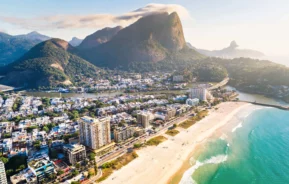 Imagem da vista aérea da paisagem da Barra da Tijuca mostra mar, faixa de areia, prédios e construções na beira da orla, vegetação e montanhas da região para ilustrar matéria sobre as praias da Barra da Tijuca, no Rio de Janeiro