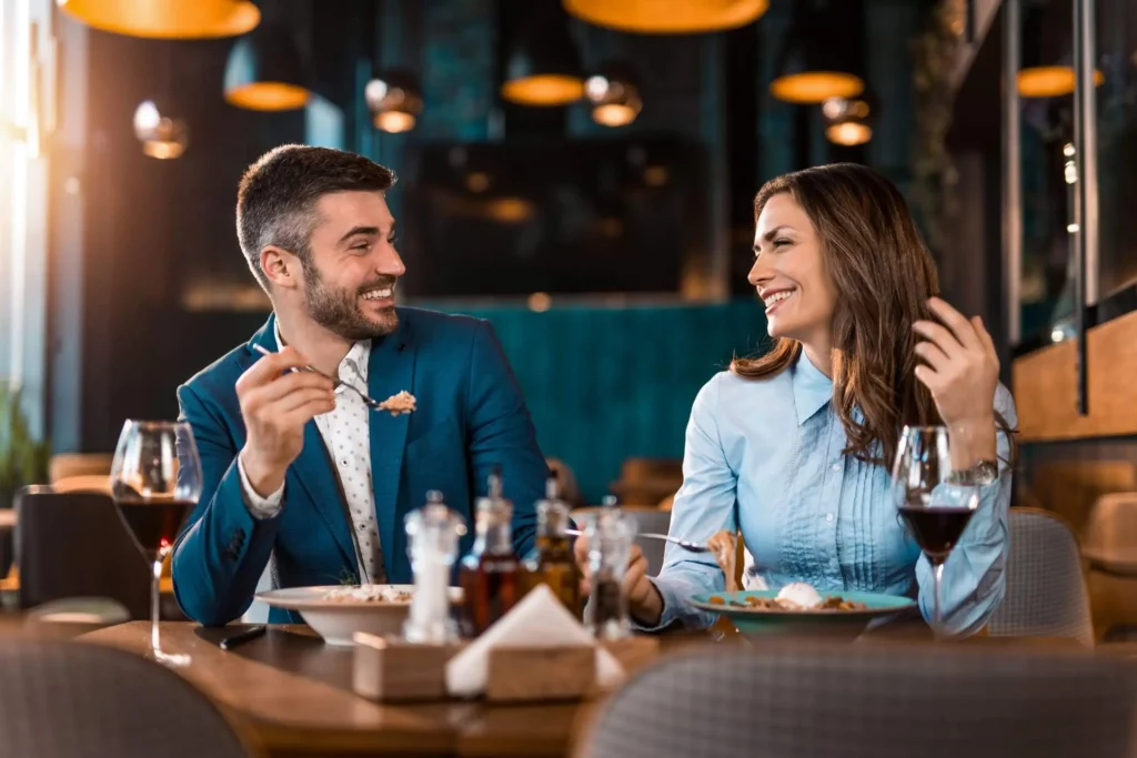 Imagem de um homem de terno azul e uma mulher de blusa azul-clara sorriem e desfrutam de uma refeição, com taças de vinho tinto sobre a mesa e uma iluminação ambiente quente ao fundo