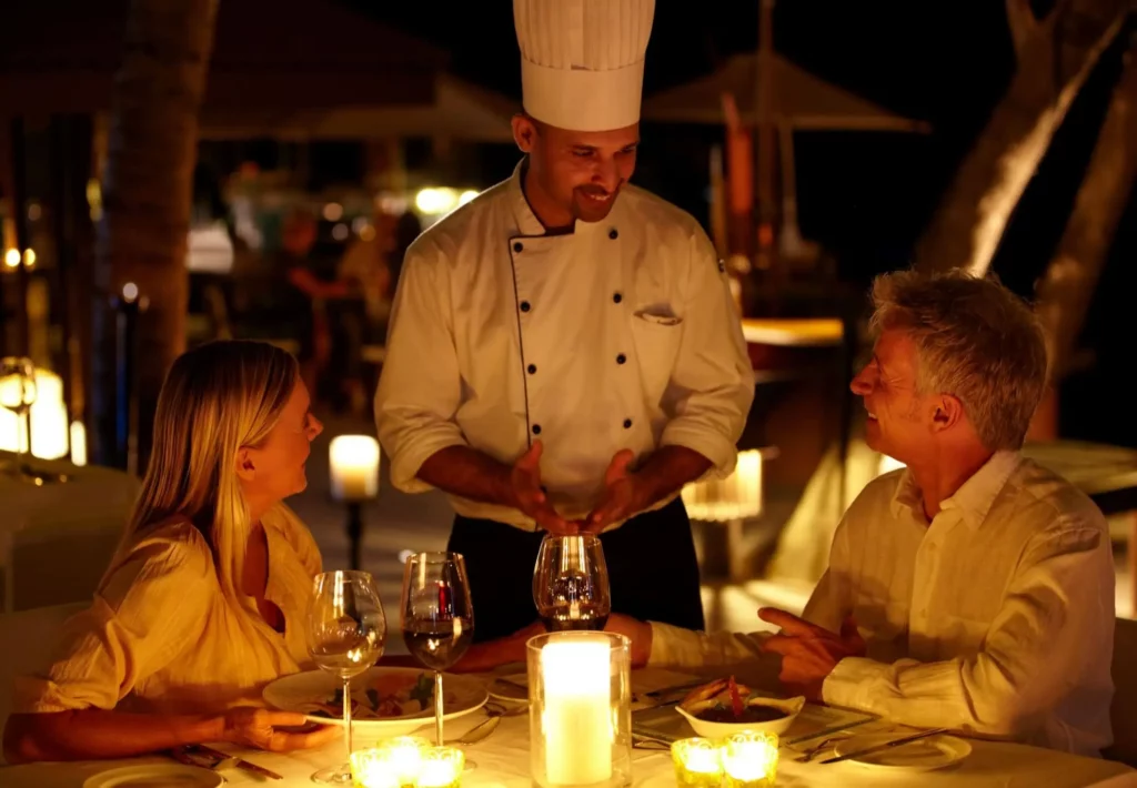 Imagem de um chef de cozinha falando com dois clientes de um restaurante que estão sentados em uma mesa iluminada por velas para ilustrar matéria sobre os restaurantes de luxo em São Paulo