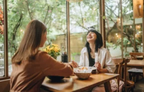 Imagem de duas mulheres sentadas em uma mesa de madeira em um restaurante aconchegante com grandes janelas, compartilhando uma refeição. Uma mulher ri alegremente enquanto a outra sorri, desfrutando da suave luz natural - apenas mais um momento agradável em um dos melhores restaurantes de São Paulo
