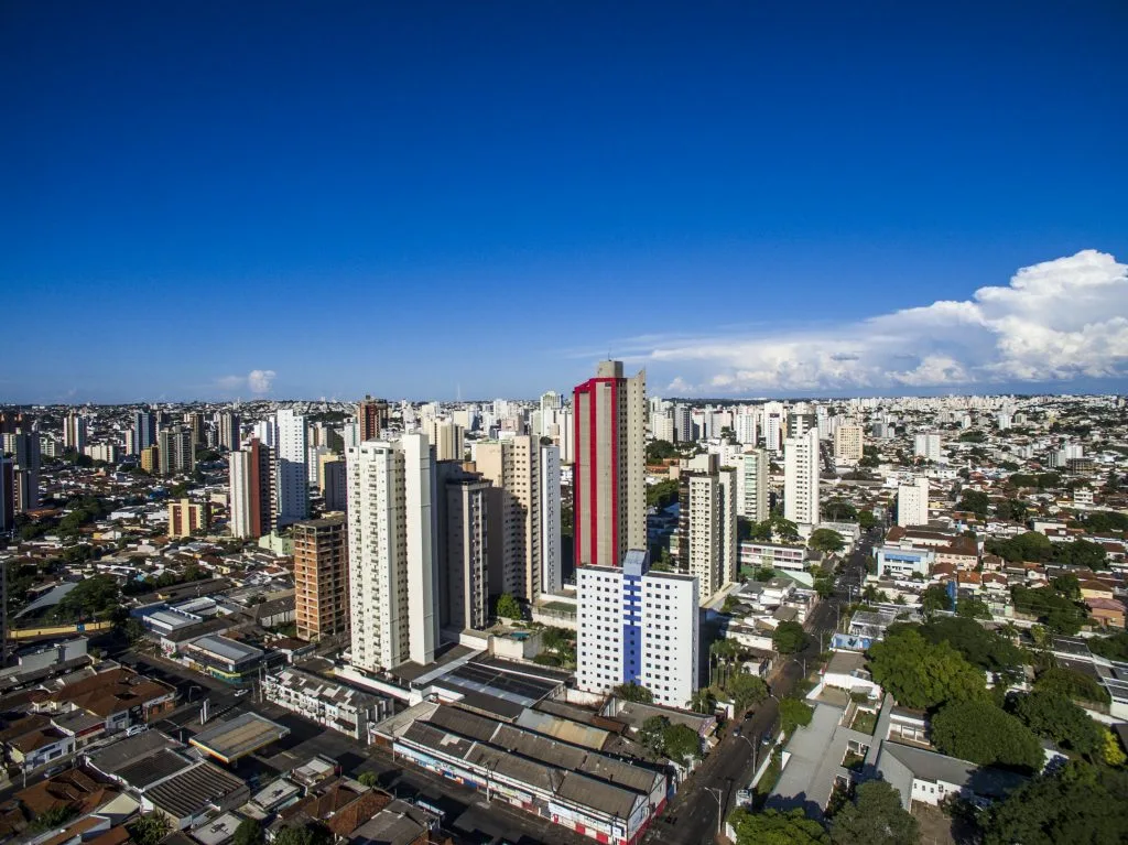 Imagem da vista aérea de Uberlândia mostra edifícios altos sob um céu azul claro, com um edifício vermelho e branco proeminente no centro, cercado por estruturas menores e vegetação