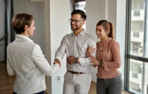 Imagem de um casal formado por um homem e uma mulher visitando um imóvel enquanto o homem aperta a mão de uma agente imobiliária para ilustrar matéria sobre apartamento Minha Casa, Minha Vida pode ser vendido?