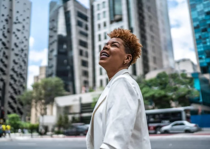 Imagem de uma mulher no centro de uma cidade cercada por prédios empresariais com um sorriso no rosto para ilustrar matéria sobre as cidades do futuro